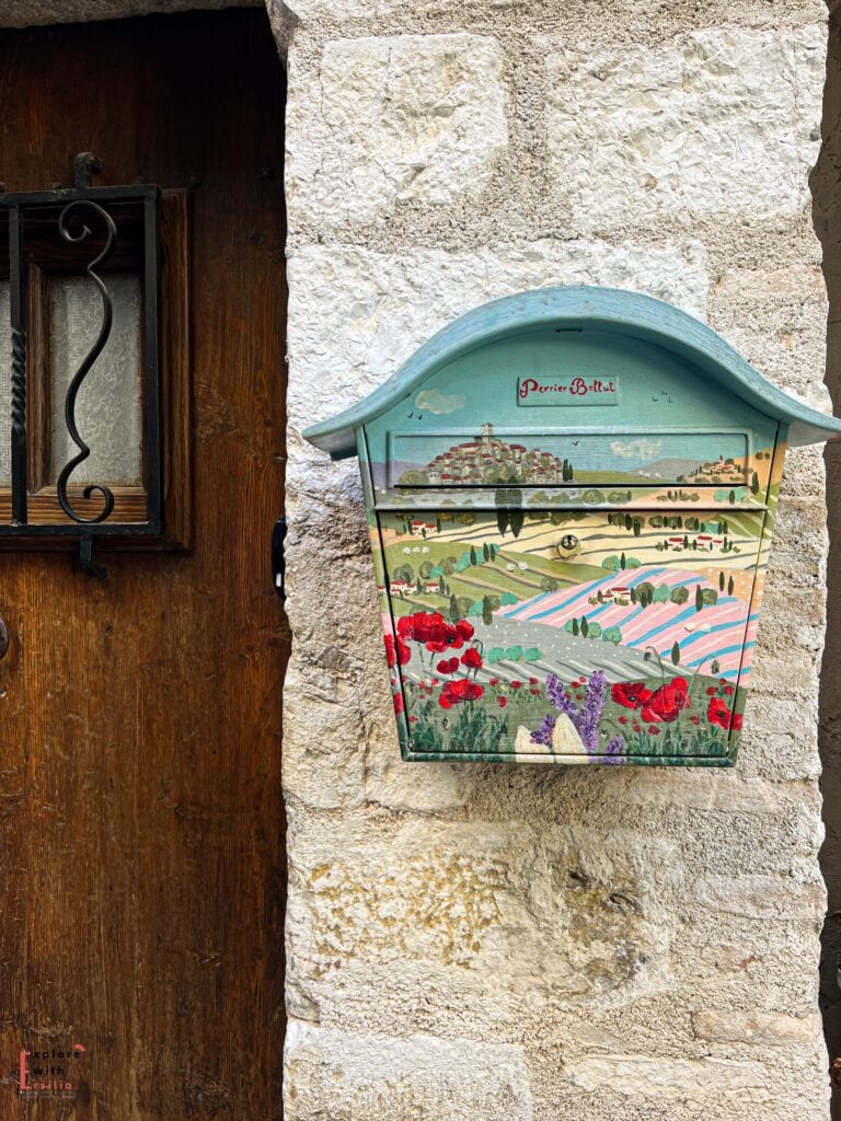 Decorative mailbox mounted on a stone wall, hand-painted with a landscape scene featuring red poppies and lavender fields in pastel colors. The mint green mailbox is framed by a wooden door with wrought iron details