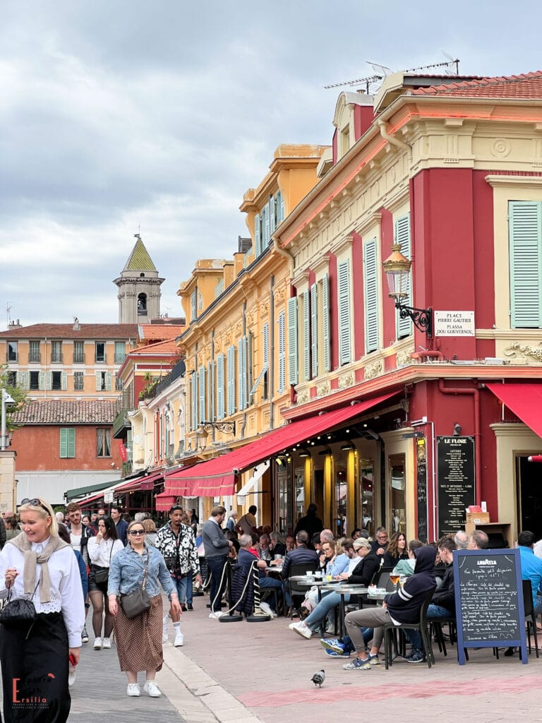 A bustling street scene in Nice's old town, with colorful buildings featuring yellow and red facades and blue shutters. Restaurants with red awnings line the street, their outdoor tables filled with diners. A church bell tower rises in the background, and pedestrians stroll along the lively thoroughfare. Chalkboard menus and vintage-style street lamps add to the traditional French atmosphere.