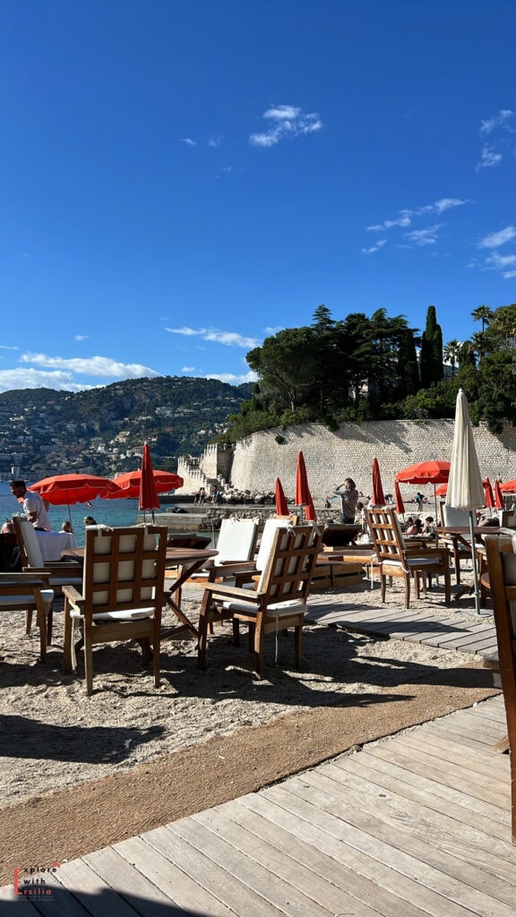 Beach club at Cap Ferrat featuring wooden lounge chairs and red umbrellas along the Mediterranean shore. Stone retaining walls topped with cypress trees rise behind the beach, while coastal mountains form the backdrop against a vivid blue sky.