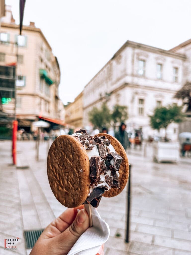 Close-up of a stracciatella ice cream sandwich with chocolate chunks held between two chocolate chip cookies against a blurred background of Nice's Old Town with historic buildings and pedestrian streets.