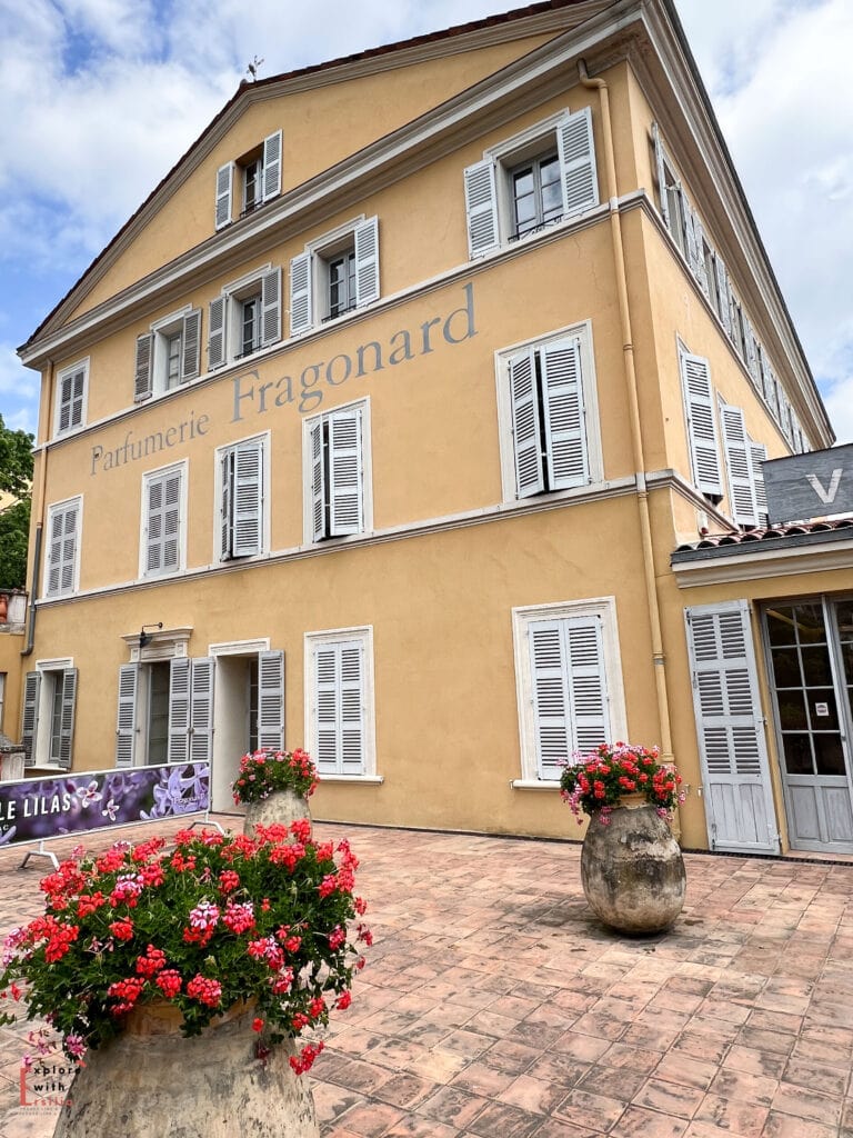 Historic Parfumerie Fragonard building in Grasse, featuring a pale yellow façade with white shuttered windows. Large stone planters with vibrant red geraniums decorate the entrance courtyard, and a lilac-colored banner is visible near the doorway. The traditional Provençal architecture includes multiple floors of symmetrical windows and a sloped roof against a blue sky.