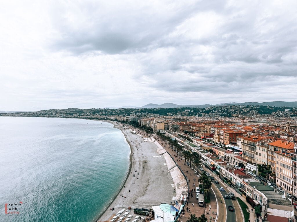 Aerial view of Nice's famous Promenade des Anglais, showing the curved coastline with its pebble beach and turquoise Mediterranean waters. The iconic palm tree-lined promenade stretches along the bay, bordered by Belle Époque architecture with terracotta roofs. The city extends into the hills behind, with the beach dotted with sunbathers and beach umbrellas under an overcast sky.