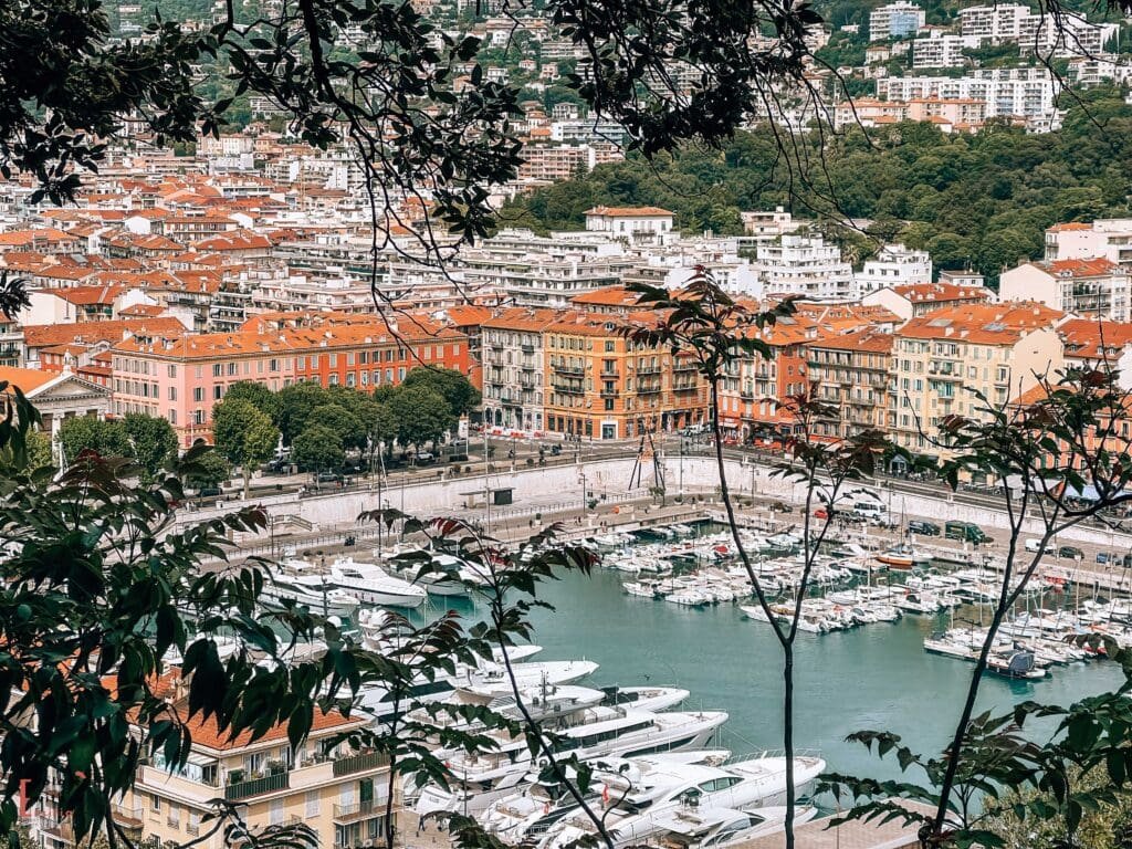 Elevated view of Port Lympia in Nice framed by tree branches, showing the harbor filled with white yachts and boats, surrounded by colorful Italianate buildings with terracotta rooftops in shades of pink, orange, and yellow, with the hillside neighborhoods of Nice rising in the background against the green hills. One of the best things to do in Nice in one day.