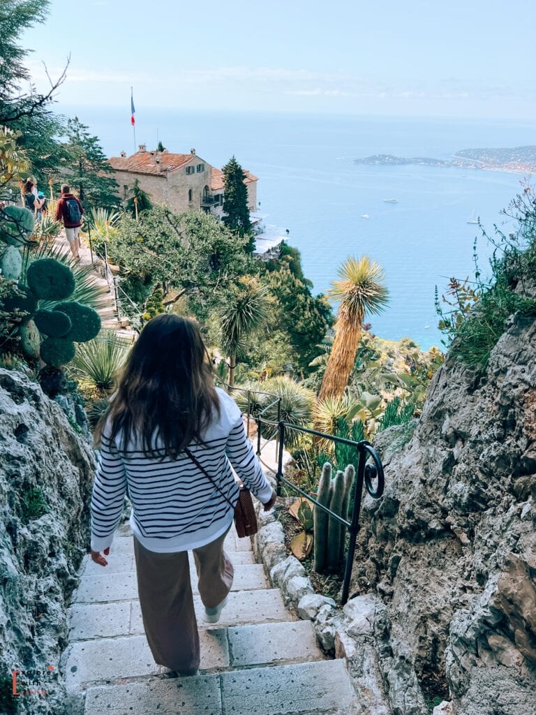 Stone steps wind through Eze's exotic garden, with a visitor in a striped sweater descending the path. Mediterranean views stretch to the horizon, with the French Riviera coastline visible below. The garden features cacti, yucca plants, and rocky outcrops, while a historic building with a French flag flies at the hilltop. The scene captures the dramatic elevation of this medieval village garden.