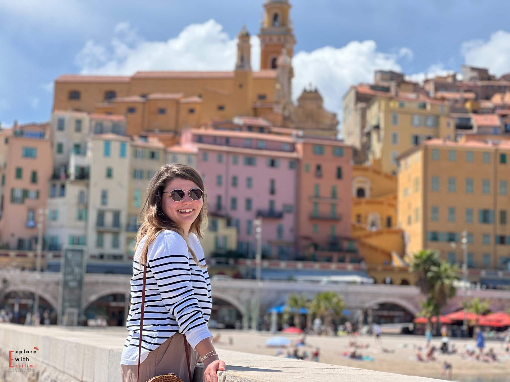 A woman wearing a striped marinière (French sailor shirt) and sunglasses smiles at the camera while standing along the beachfront in Menton, France. Behind her, the city's distinctive colorful buildings in pastel yellows, pinks, and oranges rise up the hillside, crowned by the baroque Basilica of Saint-Michel with its ochre bell towers. The beach below is dotted with sunbathers on a bright spring day.