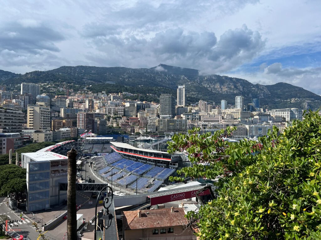 Aerial view of Monaco's iconic Formula 1 street circuit and cityscape. In the foreground, the circuit's temporary grandstands with blue seats are visible, along with a section of the race track. The city's dense urban landscape of high-rise buildings stretches up the mountainside behind the track, with luxury yachts visible in the harbor. The scene is framed by lush green foliage in the foreground and dramatic mountain peaks under cloudy skies. A Casino sign and building can be spotted among the architecture.