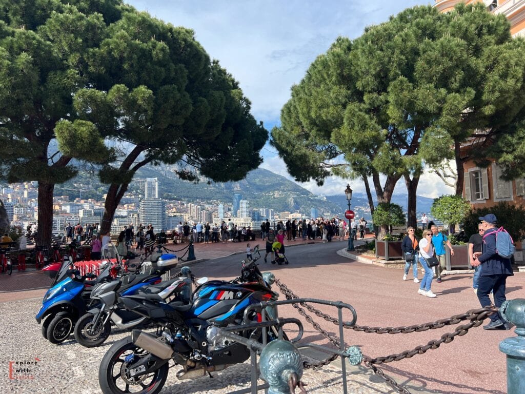 View from Monaco's scenic overlook, featuring parked motorcycles including BMW and other sport bikes in the foreground. Large umbrella pine trees frame the vista of Monaco's skyline with its modern high-rise buildings against the backdrop of mountains. Tourists gather at the viewpoint, and a vintage-style lamppost adds to the Mediterranean atmosphere.