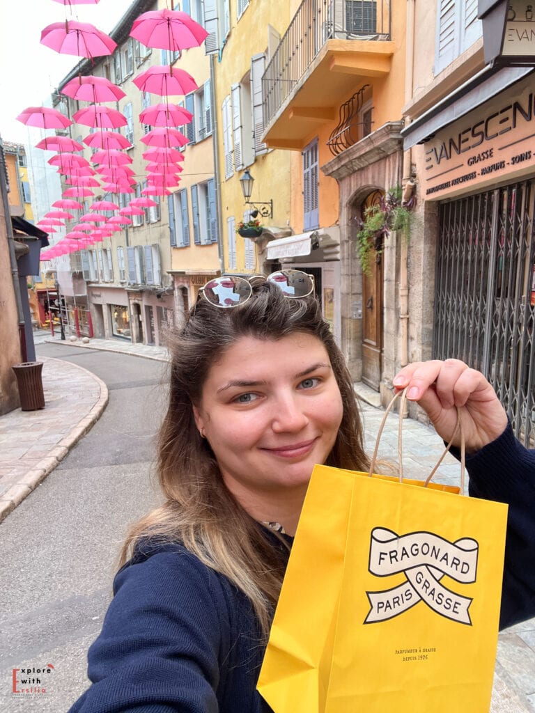 A shopping scene in Grasse showing a yellow Fragonard shopping bag being held up against a backdrop of pink umbrella art installation. The picturesque street features traditional Provençal buildings with blue-grey shutters and stone archways. Taken as a selfie in front of 'Evanescen' shop sign.