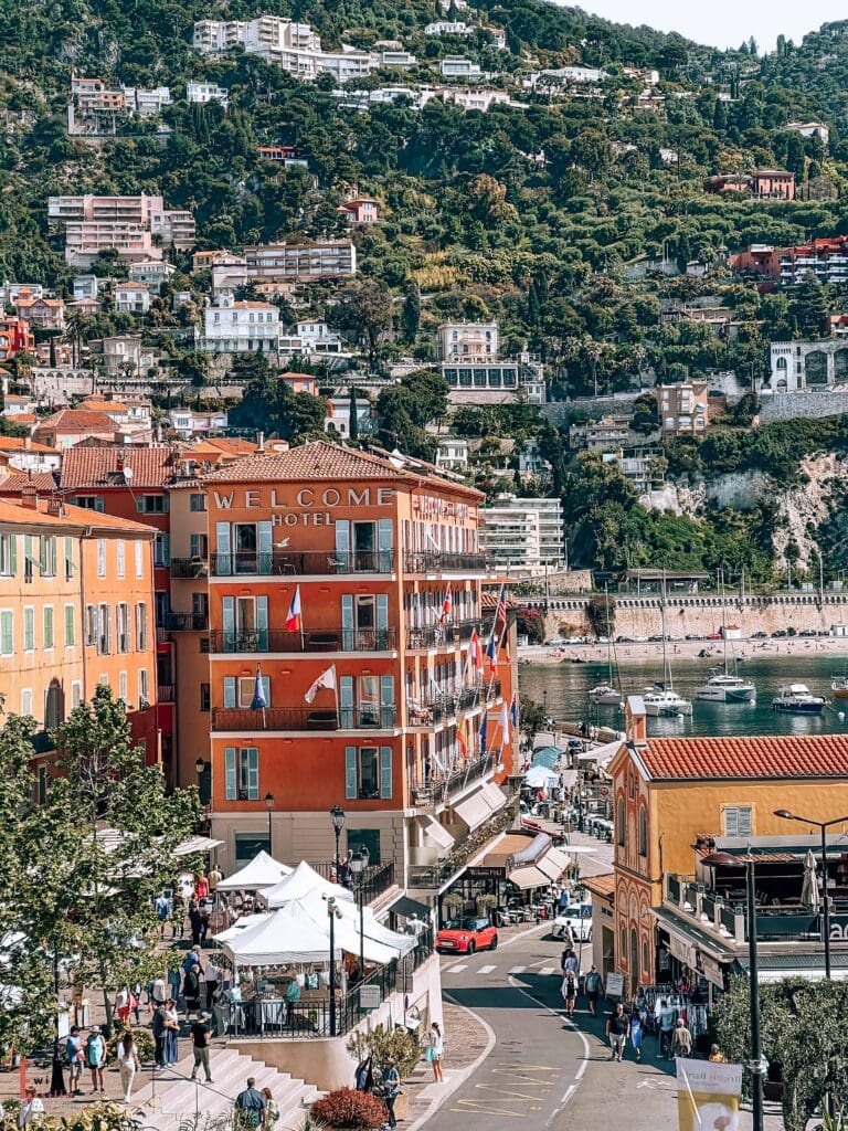 View of Villefranche-sur-Mer's waterfront, with the orange-colored Welcome Hotel as the centerpiece. The hillside is covered with terraced villas and apartments ascending the steep terrain. Market stalls with white canopies line the street below, while boats are moored in the harbor. The scene captures the typical French Riviera architecture with terracotta roofs and pastel-colored buildings against the Mediterranean coastline.