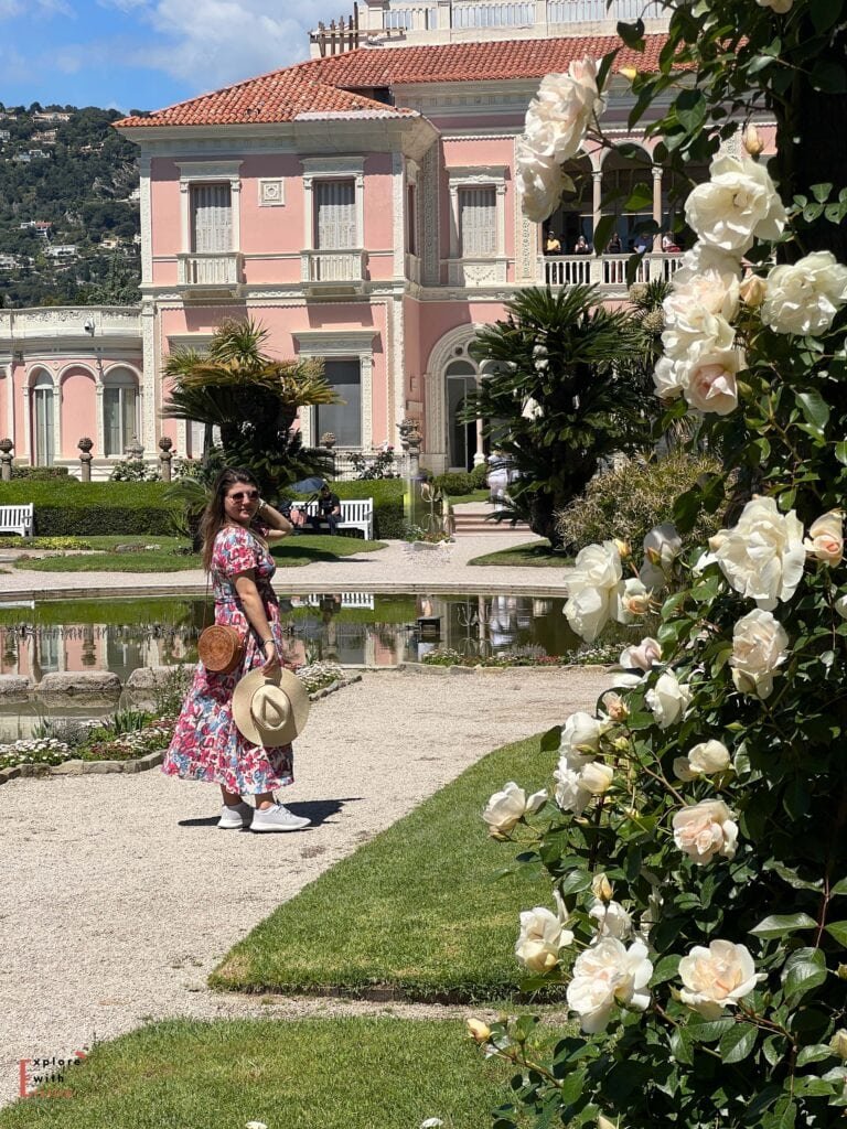  A woman in a floral dress stands in the gardens of Villa Ephrussi de Rothschild in Saint-Jean-Cap-Ferrat. The magnificent pink villa with its Italian-style architecture forms the backdrop, while white roses frame the foreground. A reflecting pool and manicured gardens with gravel paths complete the elegant scene. The woman holds a straw hat and round woven bag, standing near the ornamental pond in front of the historic mansion.