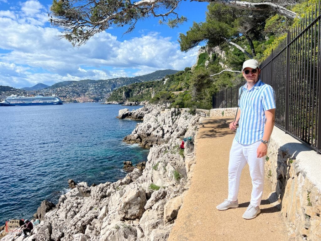 A man in striped blue and white shirt, white pants, and a baseball cap - stands on the Sentier du Littoral coastal path in Cap d'Ail. Behind him, the Mediterranean coastline stretches toward Monaco, where a large cruise ship is visible in the harbor. The path hugs dramatic limestone cliffs above turquoise waters, with the mountains of the French Riviera rising in the background under a bright blue sky with scattered clouds.