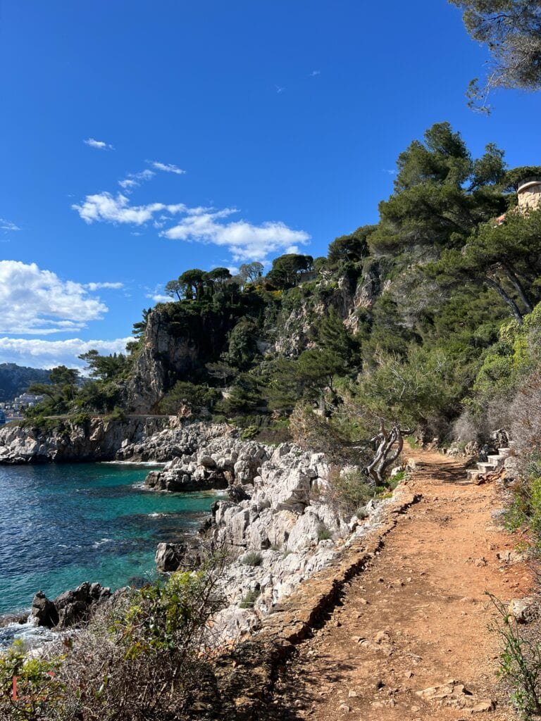 A scenic view of the Sentier du Littoral coastal path between Cap d'Ail and Monaco. The dirt trail winds along dramatic white limestone cliffs, with turquoise Mediterranean waters below and stone pine trees crowning the rocky outcrop above. The coastline curves into the distance beneath a vivid blue sky dotted with white clouds, showcasing the rugged natural beauty of the French Riviera.