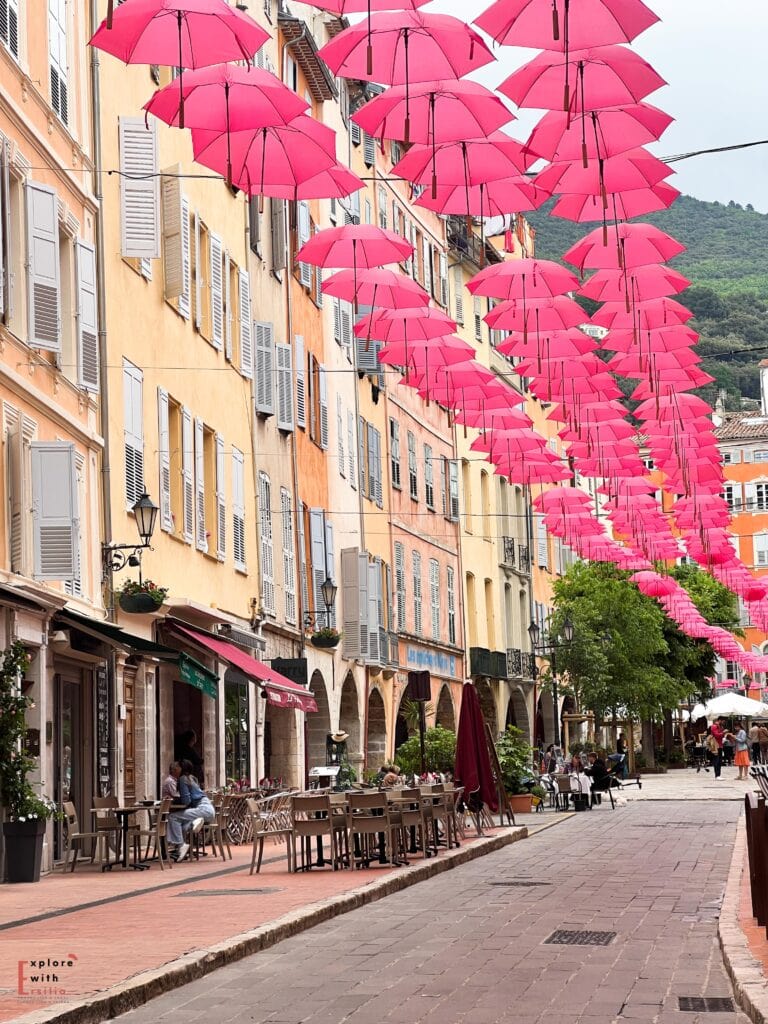 A charming pedestrian street in Grasse, French Riviera, with vibrant pink umbrellas suspended overhead. The street features traditional Provençal architecture with peach-colored buildings, grey shutters, and arched walkways. Café tables and chairs line the street beneath the umbrellas, with scattered patrons enjoying outdoor dining. Vintage-style lampposts and potted plants add to the Mediterranean atmosphere.