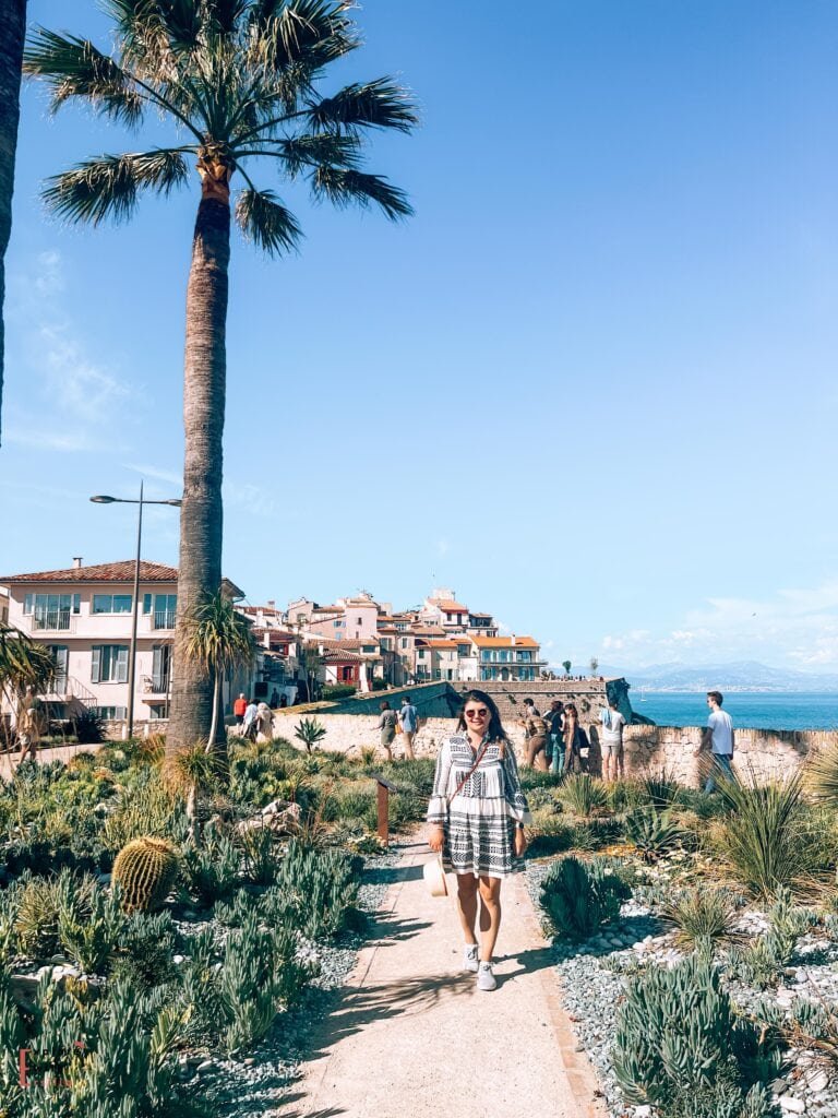 A woman in a white and black patterned dress walks along a Mediterranean garden path in Antibes. The landscaping features drought-resistant plants including tall palm trees, barrel cacti, and coastal vegetation. Behind her, the historic ramparts and pastel-colored buildings of old Antibes rise up, with the blue Mediterranean Sea and mountains visible in the distance. The scene captures the essence of the French Riviera with its mix of cultivated gardens, historic architecture, and seaside views.