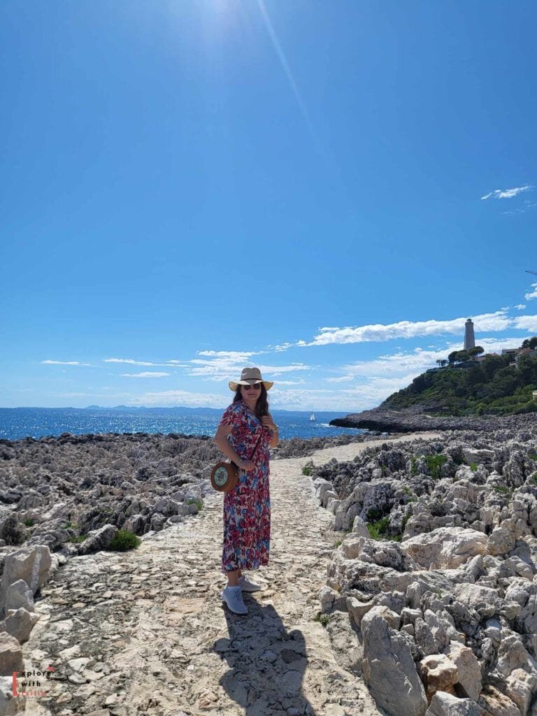 A woman in a floral dress and sunhat stands on a rocky coastal path at Cap Ferrat, with the lighthouse of Saint-Jean-Cap-Ferrat visible on the hilltop in the background. The Mediterranean Sea sparkles a brilliant blue on this sunny day, with a small sailboat visible on the horizon. The path winds through limestone rocks typical of the French Riviera coastline, creating a rugged yet scenic landscape.