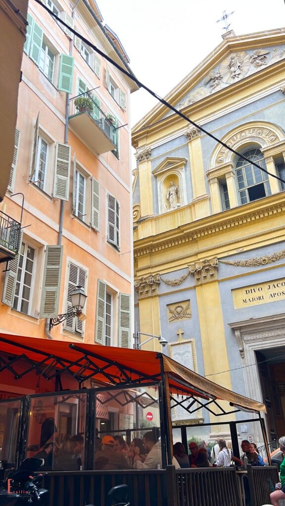 View of Saint-Jacques-le-Majeur Church in Nice featuring its ornate yellow and pale blue Baroque façade with classical pediment, religious sculptures, and decorative elements, juxtaposed against a traditional coral-colored residential building with green shutters and an outdoor café with red awning on the narrow street below.