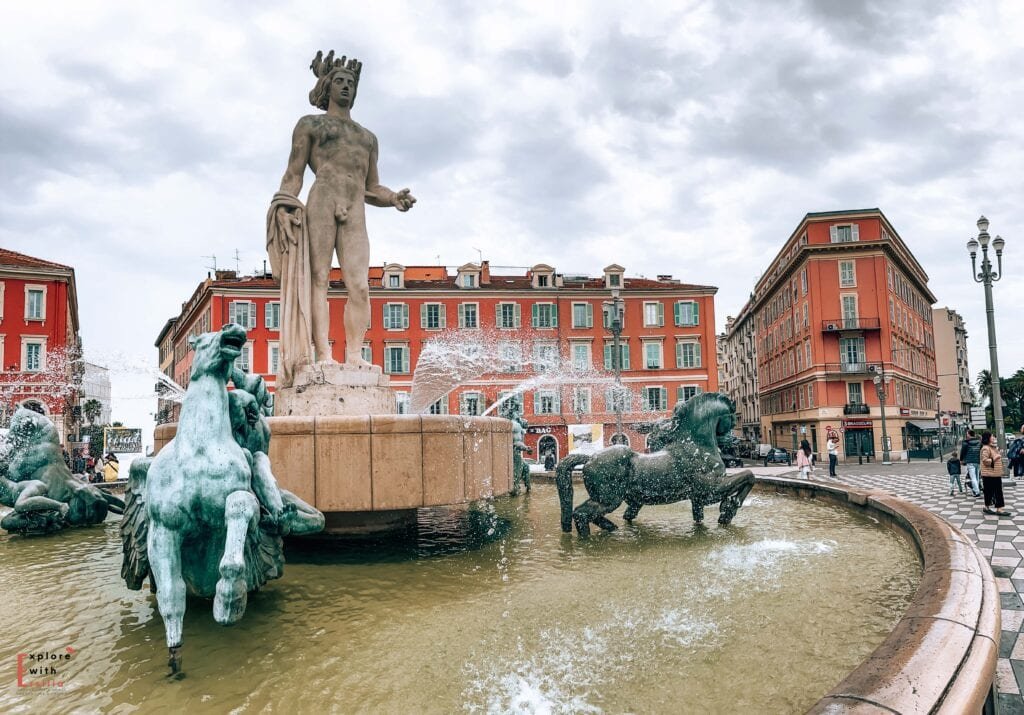 Place Masséna in Nice, featuring the Apollo Fountain at its center with a tall crowned statue surrounded by bronze horses emerging from the water. The fountain is framed by the square's distinctive red ochre buildings with blue shutters and white trim, exemplifying the Italian-influenced Sardinian architecture. The geometric patterned pavement and ornate lampposts complete this main square's classical Mediterranean design.