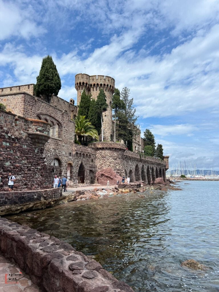 The Château de La Napoule rises dramatically from the Mediterranean shoreline, photographed in spring 2024. Its medieval stone walls, crenellated tower, and arched sea wall stretch along the water's edge. The reddish stone of the fortress contrasts with tall cypress trees and a palm tree on its grounds. In the background, sailboats are moored in the harbor, and tourists walk along the sea wall path. A bright blue sky with wispy clouds completes the scene of this French Riviera landmark.