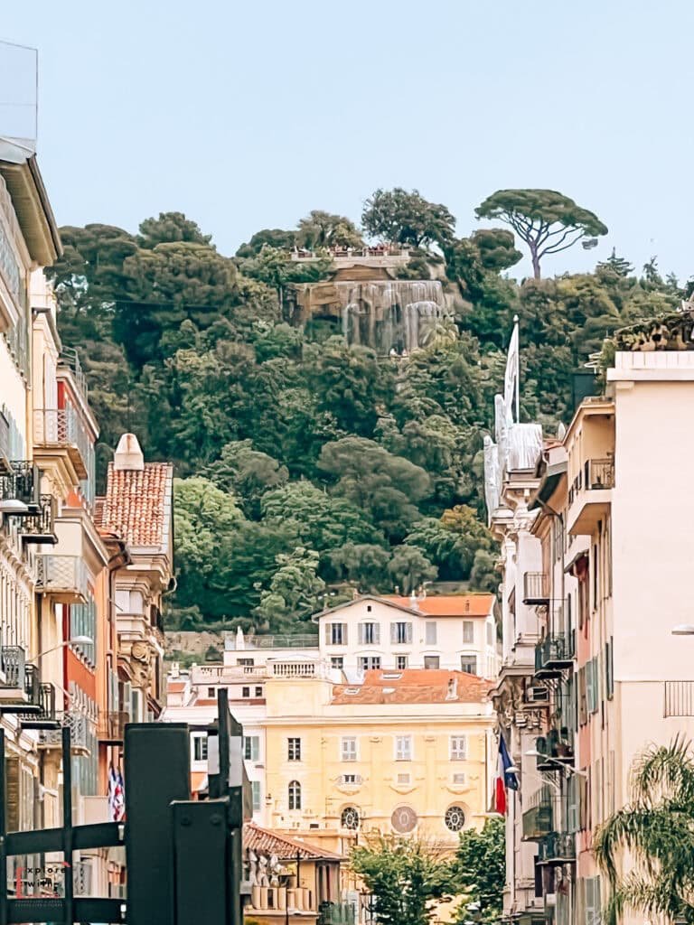 The Colline du Château seen from the city with the waterfall beign visible on the top.  Colored buildings on the streets in the city. 