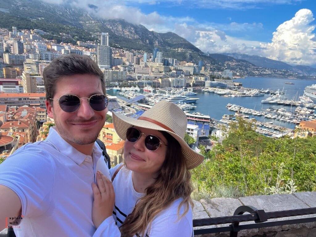 "Selfie overlooking Monaco's Port Hercule, with a couple in matching white outfits and sunglasses. The backdrop showcases Monaco's luxury yacht marina, high-rise buildings climbing the mountainside, and the Mediterranean coastline. A straw hat and clear sunny skies complete the summer scene.