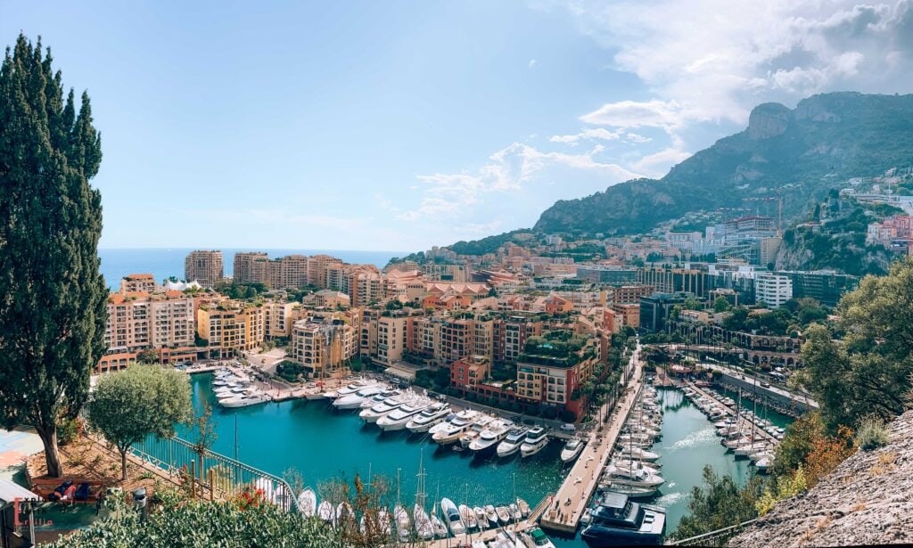 Panoramic view of Monaco's Fontvieille district showcasing the luxury yacht-filled harbor with turquoise waters, surrounded by terracotta-roofed residential buildings, against a backdrop of dramatic Mediterranean mountains, with cypress trees framing the foreground and the azure sea visible on the horizon.