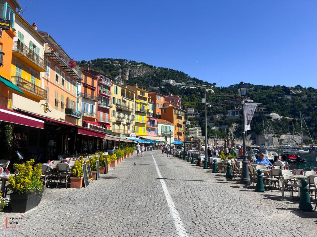 Villefranche-sur-Mer's waterfront promenade lined with vibrant multi-colored buildings in yellow, orange, and pink hues. Restaurant terraces with awnings and potted flowers stretch along the cobblestone walkway, with diners enjoying outdoor seating by the yacht-filled harbor. The hills of the French Riviera rise in the background under a bright blue sky.