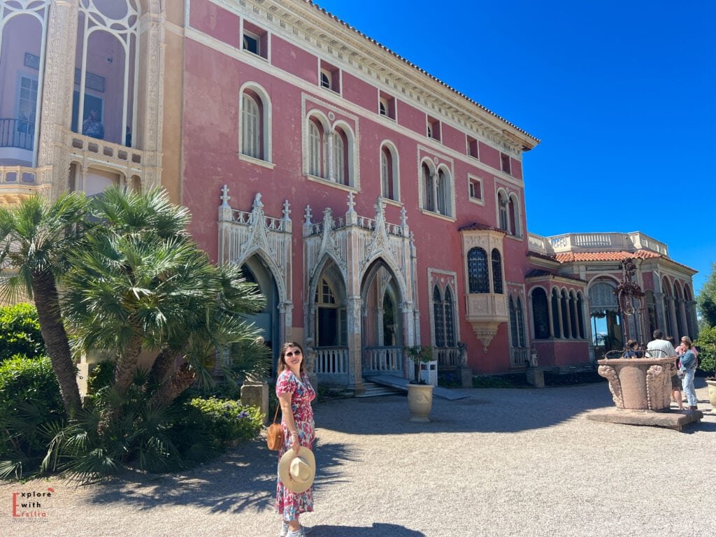 Woman in a floral dress and straw hat standing in front of Villa Ephrussi de Rothschild's Gothic-inspired entrance, with its distinctive pink façade and ornate white stone archways. The decorative architecture features pointed arches, delicate carved details, and palm trees framing the grand doorway.