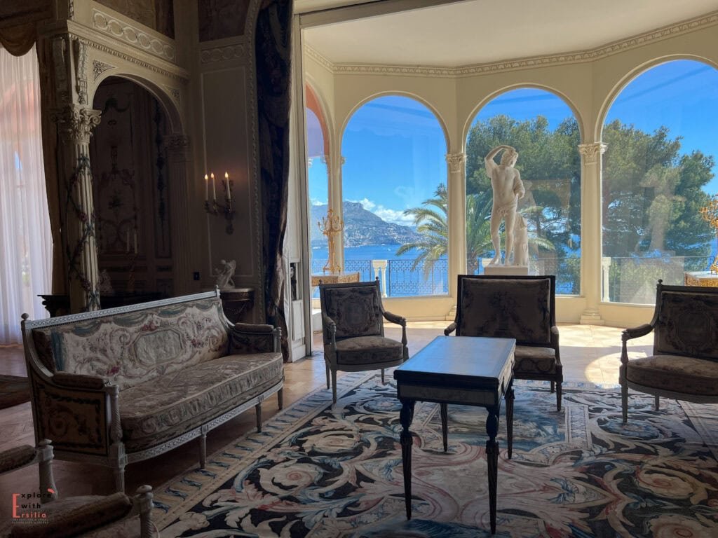 Interior of Villa Ephrussi de Rothschild's grand salon, featuring period furniture including an ornate settee and velvet armchairs arranged around a dark coffee table. Arched windows frame Mediterranean views and palm trees, with a classical statue silhouetted against the sea. Crystal candelabras and elaborate moldings complete the Belle Époque décor.