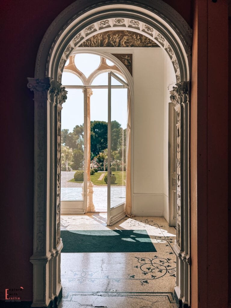 Interior archway at Villa Ephrussi de Rothschild, featuring ornate carved details and decorative columns. The doorway frames a bright view of the gardens, with sunlight streaming across the marble floor and patterned carpet. The architecture showcases the villa's elegant Belle Époque design with classical elements.