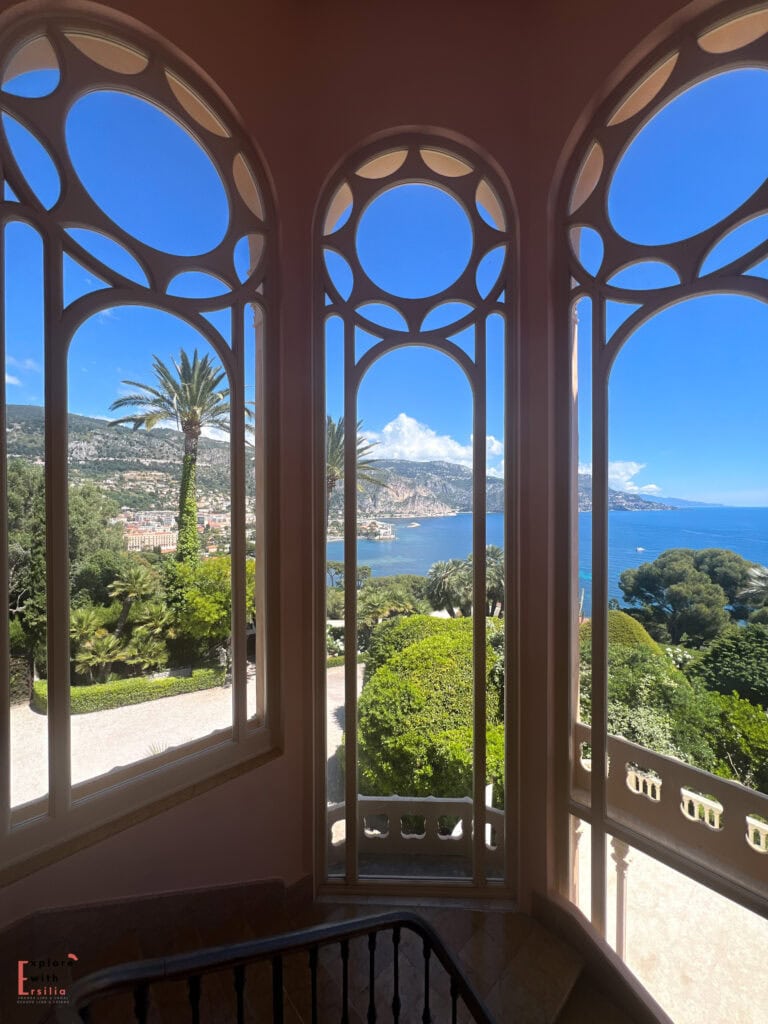 View from a decorative alcove at Villa Ephrussi de Rothschild, framed by ornate circular window patterns. The vista overlooks the Mediterranean coastline and gardens, with palm trees and manicured hedges in the foreground and the French Riviera's mountains stretching along the bay under a bright blue sky.