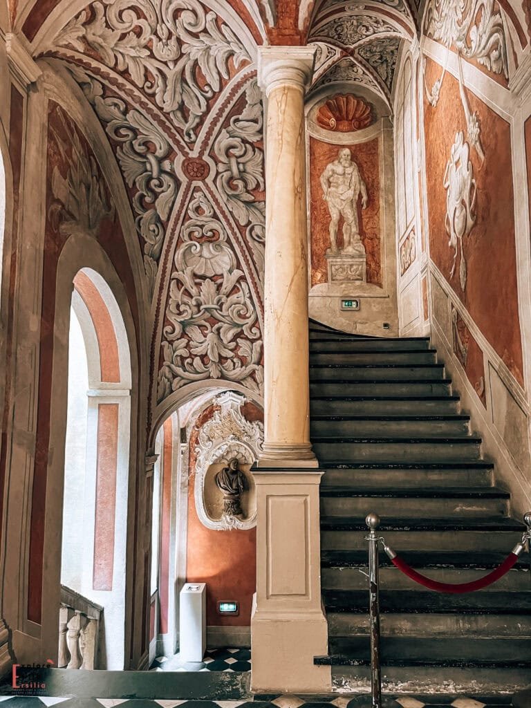 Interior staircase of Palais Lascaris in Nice, showcasing ornate Baroque architecture with elaborate stucco decorations and frescoes. A marble column stands against intricately carved white relief work on terracotta walls. Classical statuary sits in a wall niche above the stairs, and the vaulted ceiling features detailed scrollwork patterns. A modern exit sign provides a subtle contemporary contrast to the 17th-century palatial design.