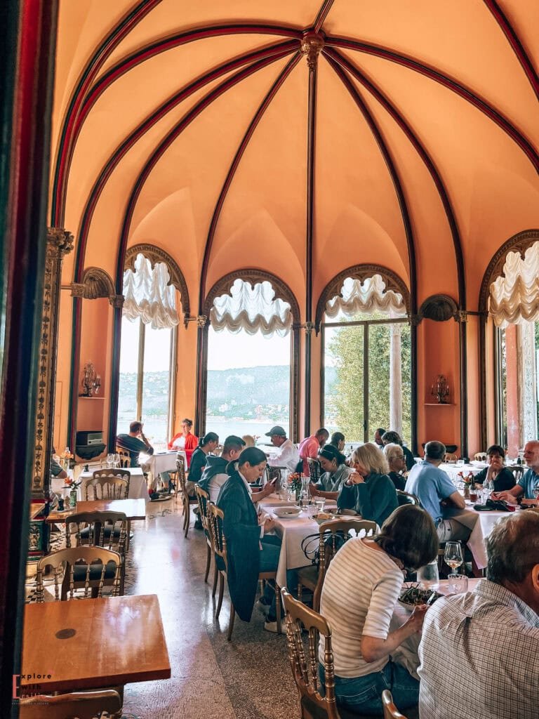 Interior of the Villa Ephrussi de Rothschild's tea room, featuring a peach-colored domed ceiling with decorative arches and large windows overlooking the Mediterranean gardens. Visitors dine at bistro tables under the elegant Belle Époque architecture of this historic villa's Salon de thé de Beatrice.