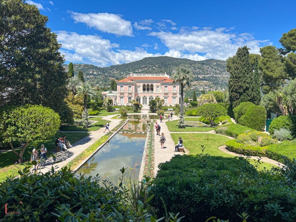 Villa Ephrussi de Rothschild's pink façade and formal gardens, featuring a long reflecting pool lined with statues and manicured lawns. Visitors stroll along the garden paths, with the villa's Italian Renaissance-style architecture set against the backdrop of Mediterranean hills and blue skies.