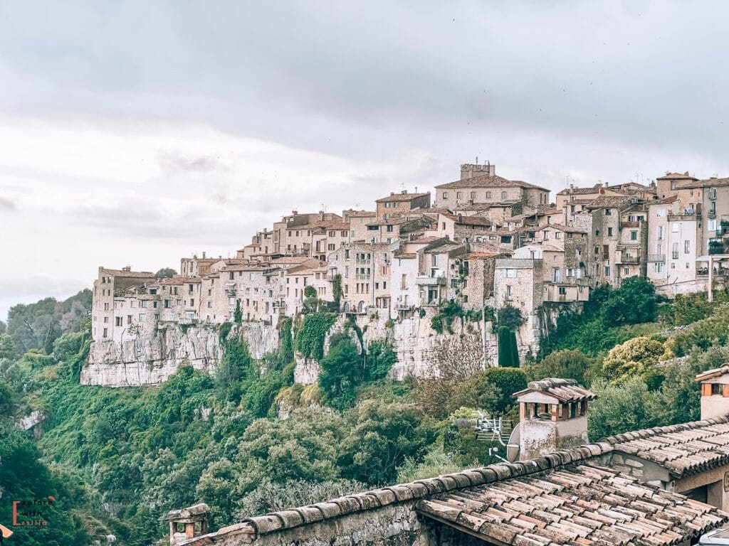 The medieval village of Tourrettes-sur-Loup dramatically perched on a cliff edge in the French Riviera. Stone houses with terracotta roofs are stacked vertically along the limestone escarpment, creating a distinctive wall-like facade. In the foreground, traditional tiled rooftops and chimneys are visible, while lush green vegetation covers the lower slopes of the cliff. The village's architecture blends seamlessly with the natural rock face, exemplifying the characteristic layout of a Provençal perched village.