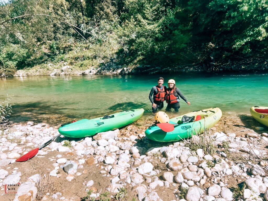 A couple wearing life jackets and safety helmets stands in emerald-green water beside their green kayaks on a rocky riverbank. The kayaks rest on a pebbly beach, surrounded by lush vegetation. The water has a striking turquoise color characteristic of the Verdon Gorge, with the deep green forest creating a natural backdrop for this kayaking adventure.