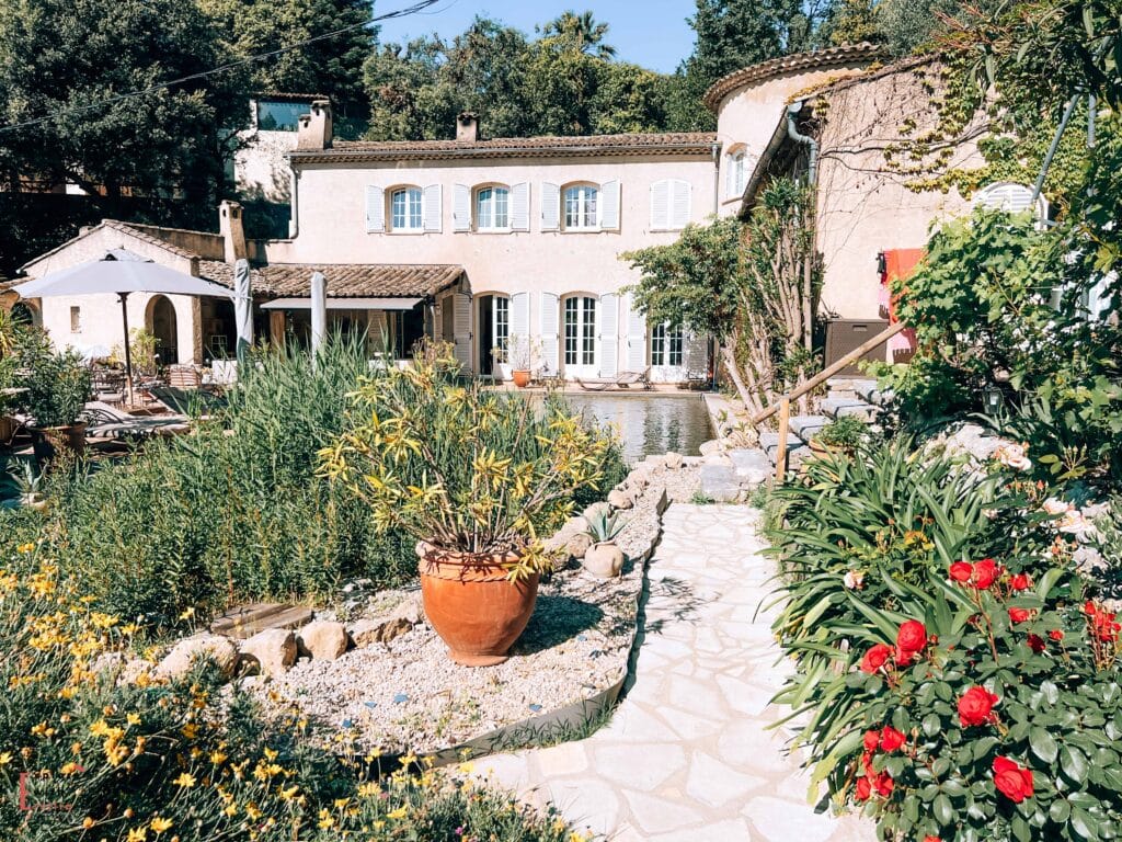 A charming Provençal bed and breakfast, Le Mazet de Tourrettes, featuring a traditional cream-colored building with pale blue shutters and terracotta roof tiles. The garden path leads through a Mediterranean landscape with yellow daylilies, red roses, and decorative grasses surrounding a natural-style pool. Large terracotta pots and stone accents complete the landscaping, creating a welcoming and tranquil setting typical of the French Riviera.