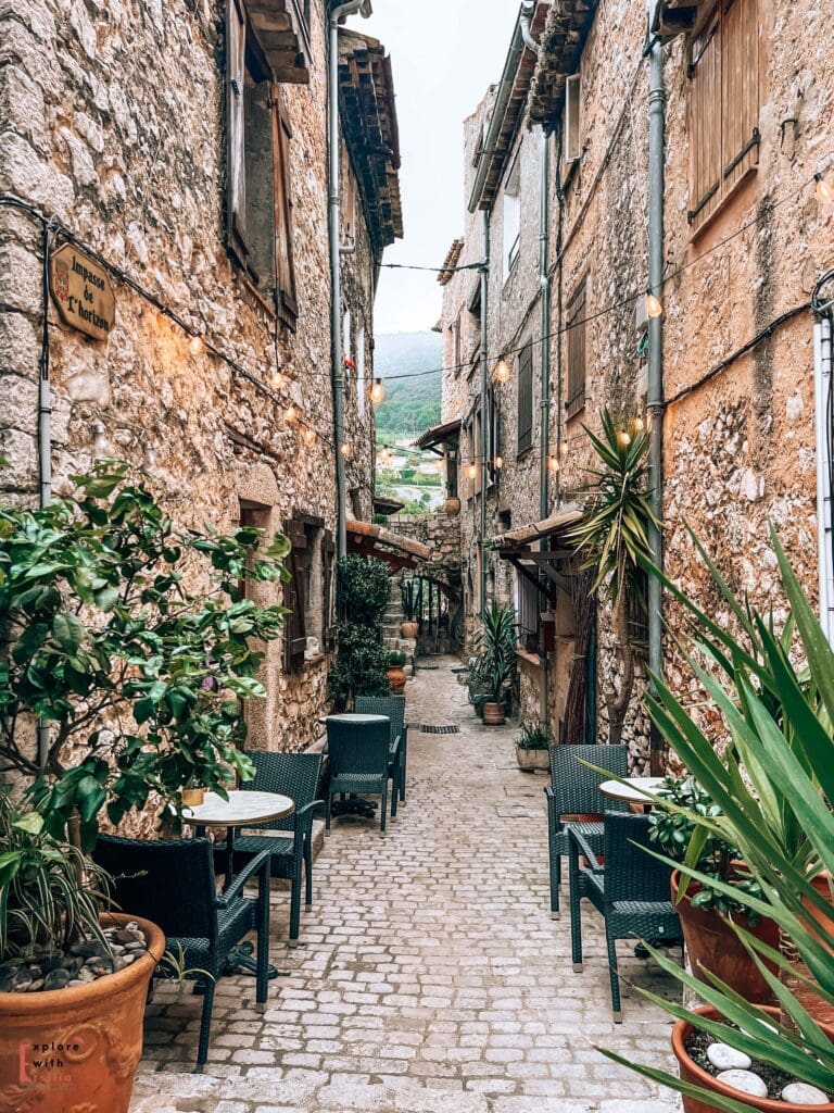 A narrow cobblestone alley in Tourrettes-sur-Loup with outdoor café seating. Dark wicker chairs and small round tables line the passage, which is decorated with string lights and potted plants including yucca and citrus trees in terracotta pots. The medieval stone walls rise high on both sides, with glimpses of the surrounding hills visible at the end of the alley. The scene captures the intimate atmosphere of a French village restaurant terrace.