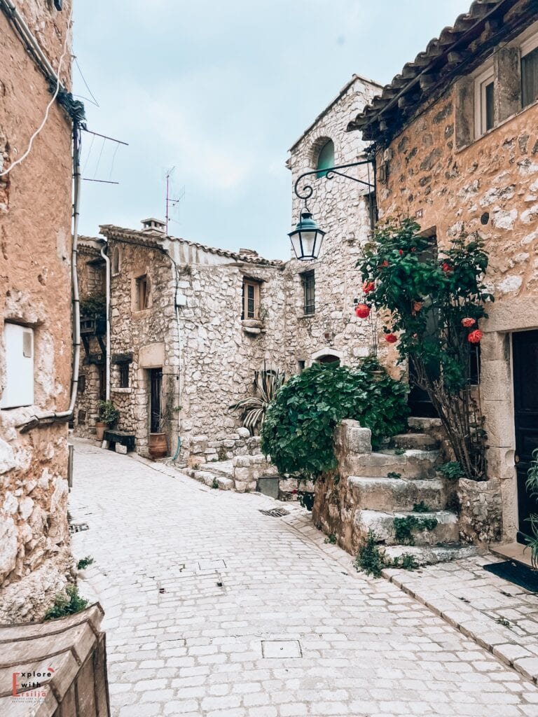 A cobblestone street winds through medieval stone buildings in Tourrettes-sur-Loup. The pale limestone walls are accented by a traditional iron street lamp and climbing roses with bright red blooms. Stone steps lead to doorways at different levels, and terracotta pots with plants add touches of greenery. The rustic scene captures the authentic character of a Provençal village street, with its weathered stones and architectural details.