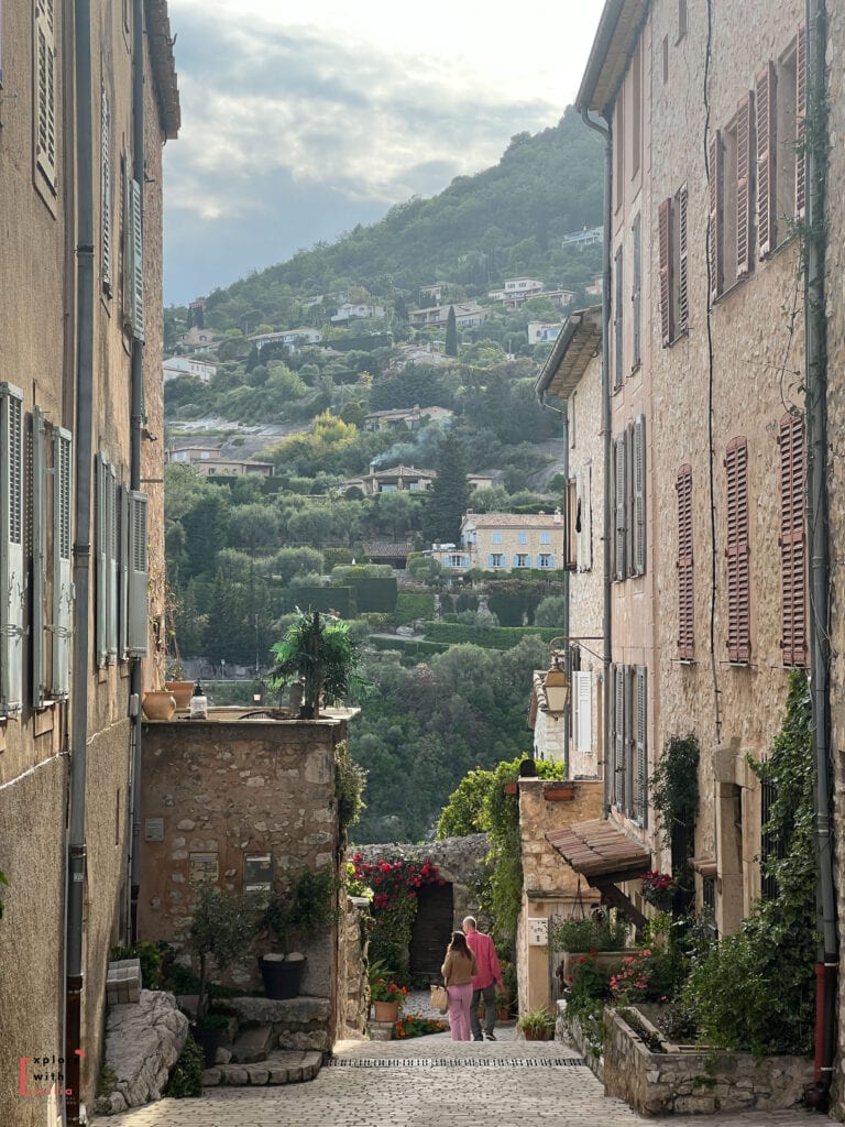 A picturesque narrow street in Tourrettes-sur-Loup, with stone buildings and shuttered windows framing a view of the valley below. A couple walks down the cobblestone path, which is decorated with potted flowers and climbing plants. Modern villas dot the green hillside in the distance, creating layers of habitation on the steep terrain. The scene captures the characteristic vertical architecture of a French Riviera hillside village, with its mix of historic stone buildings and Mediterranean vegetation.