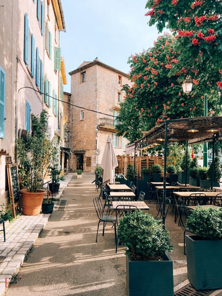 A charming street scene in Tourrettes-sur-Loup with an outdoor café terrace in the foreground. Black metal chairs and wooden tables are arranged under a rustic pergola, with green planters and potted plants creating a natural border. The stone buildings feature traditional blue shutters, and a flowering red bougainvillea tree adds a vibrant splash of color. The medieval stone tower in the background and pale limestone walls create a quintessential Provençal atmosphere.