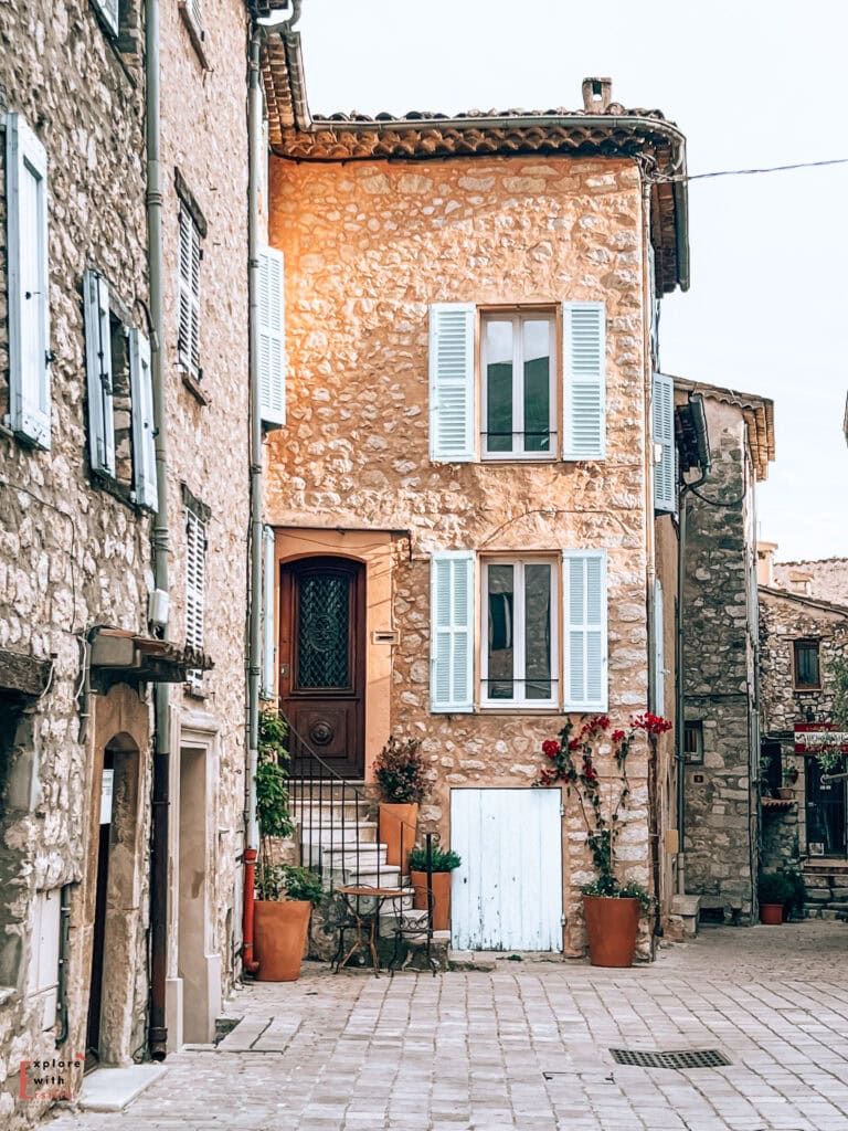 A charming stone house in Tourrettes-sur-Loup with pale blue shutters and a wooden door. The façade features traditional Provençal elements including rustic stonework, a terracotta-tiled roof, and climbing red bougainvillea. Terra cotta planters line the small entrance steps, and a white wooden garage door is set below. The narrow cobblestone street and surrounding stone buildings create an intimate village atmosphere typical of medieval French villages.