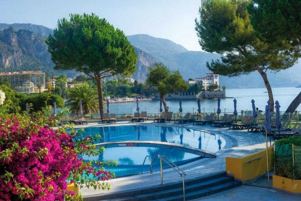 Luxury hotel pool terrace in Cap Ferrat overlooking the Mediterranean Sea. The curved swimming pool is surrounded by sun loungers and blue umbrellas, with vibrant bougainvillea in the foreground. Pine trees frame the view of the bay and mountains, while a classical villa can be seen along the waterfront.