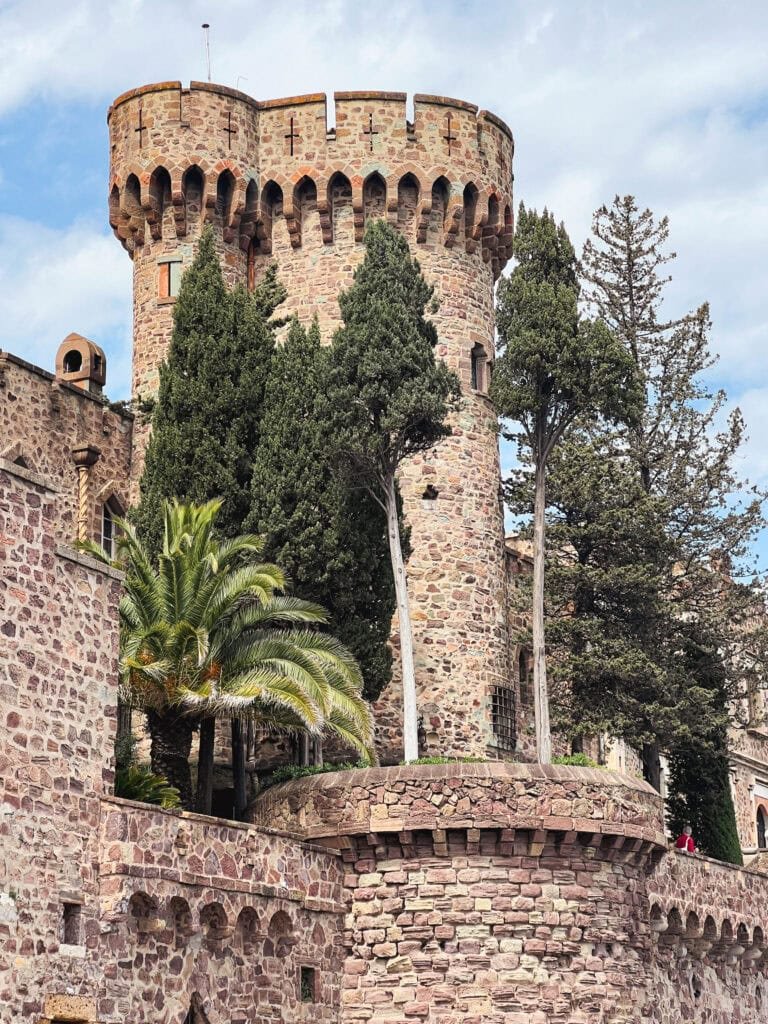 Imposing round tower of Château de la Napoule featuring crenellated battlements, multicolored stone masonry, arched windows, surrounded by tall cypress trees and palm fronds, rising above stone terraces against a blue Mediterranean sky.