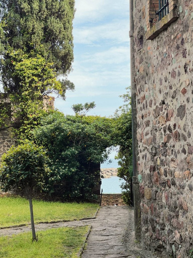 "Hidden pathway at Château de la Napoule with ancient stone walls, lush Mediterranean vegetation, and a glimpse of turquoise Mediterranean waters framed by cypress trees and shrubs, showing a secluded stone walkway leading to the sea.