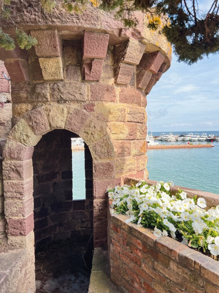 Stone passageway in Château de la Napoule's medieval tower overlooking the Mediterranean Sea and marina, featuring multicolored stonework, an arched doorway, and white petunias blooming in a brick planter box.