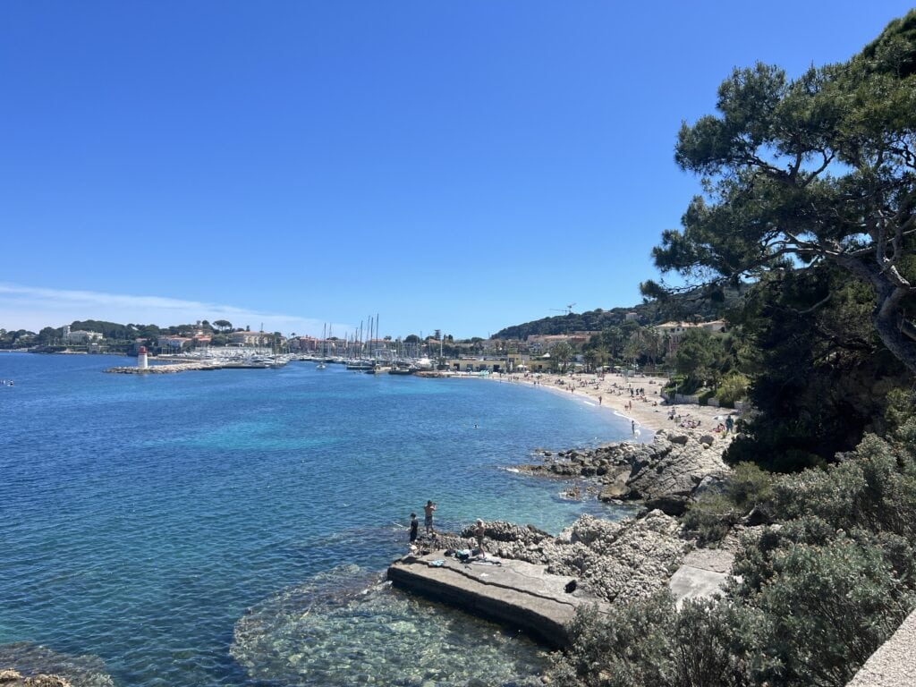 Scenic view of Saint-Jean-Cap-Ferrat's coastline showing a curved beach and marina. The crystal-clear turquoise waters meet rocky outcrops where people are swimming, with a white lighthouse visible at the harbor entrance. Pine trees frame the view of the beach dotted with sunbathers, while boats and yachts fill the marina against a backdrop of Mediterranean buildings and hills.