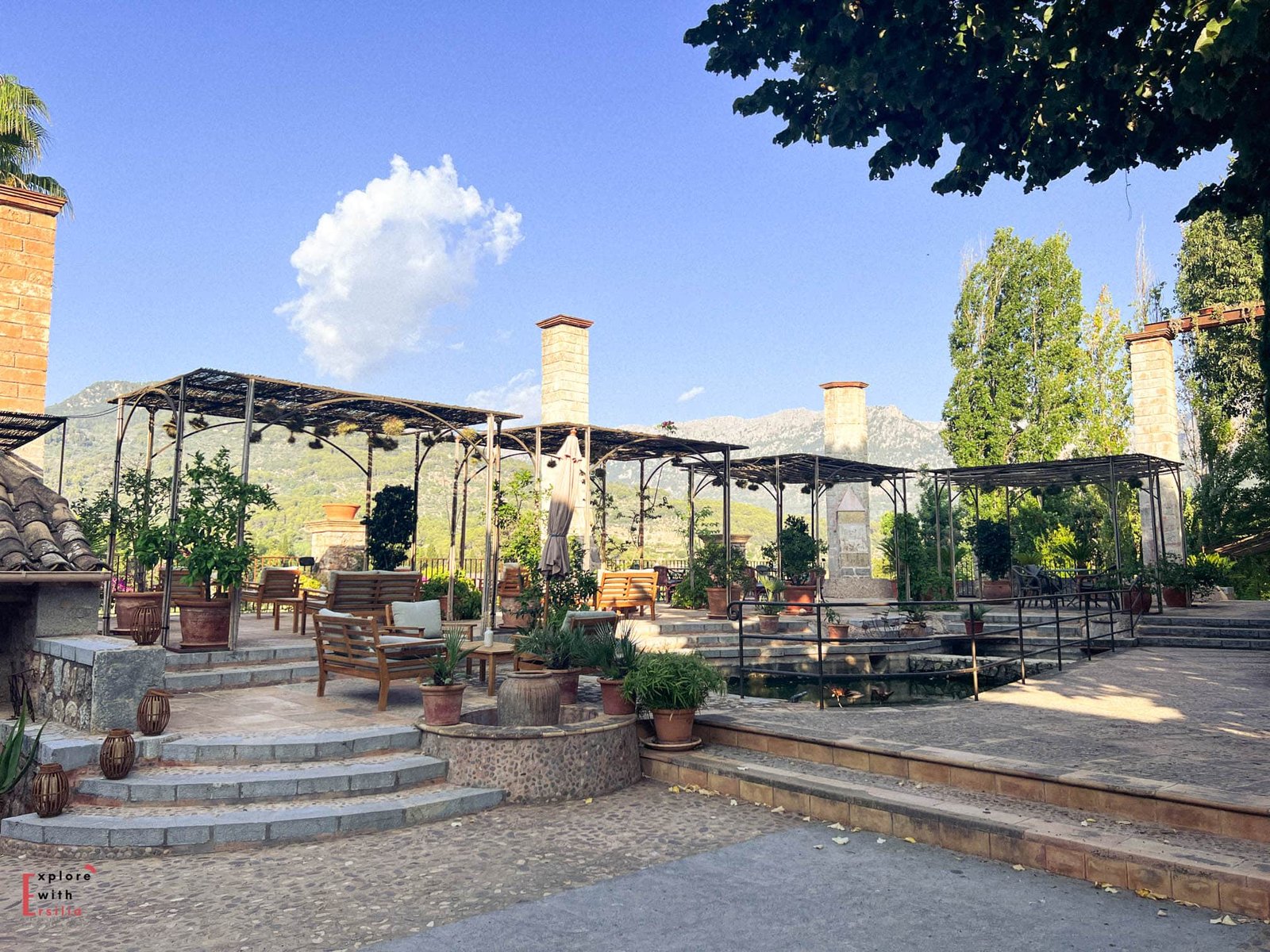 Terraced outdoor lounge area at Finca Can N'ai featuring multiple covered seating areas with wooden benches, terracotta planters, and stone steps, all set against the backdrop of the Tramuntana mountains under a bright blue sky.