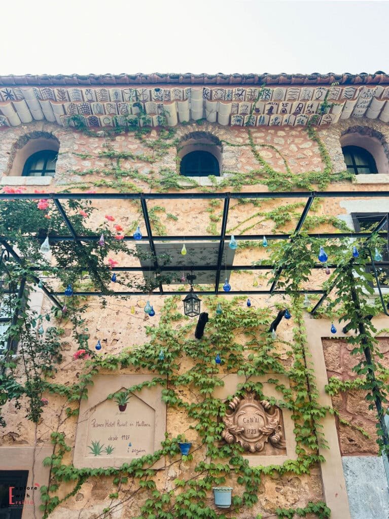 Historic facade detail of Finca Can N'ai showing decorative roof tiles with artistic patterns, climbing ivy on stone walls, blue glass pendant lights, and a carved stone plaque from 1723 alongside a painted inscription celebrating the property's heritage as a rural hotel.