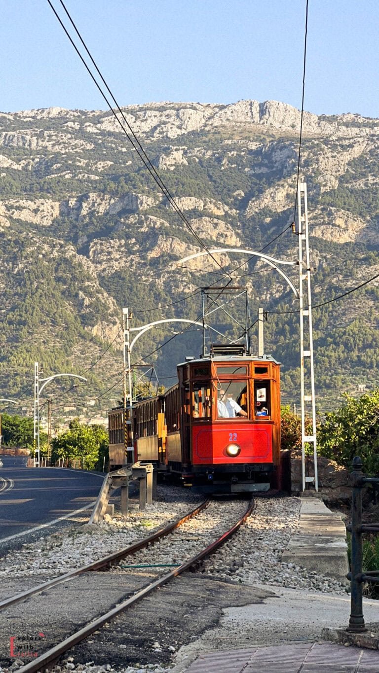 Historic red tram running on tracks between Sóller and Port de Sóller, with the dramatic limestone cliffs of Serra de Tramuntana mountains rising in the background against a clear blue sky.