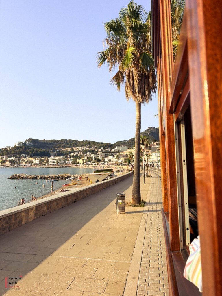 View from the vintage tram window of Port de Sóller's waterfront promenade, with palm trees lining the path, beachgoers enjoying the shoreline, and the town's buildings rising up the hillside in the golden afternoon light.